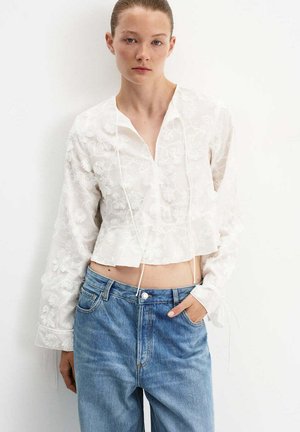 Mujer con un top corto blanco bordado de manga larga y jeans azul claro de tiro alto, de pie frente a un fondo blanco liso.