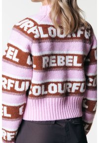 Henkilö, joka on pukeutunut pörröiseen neulepaitaan, jossa on vaaleanpunaisia, ruskeita ja valkoisia raitoja, ja jossa on sanat "REBEL" ja "COLOURFUL" lihavoinnilla.