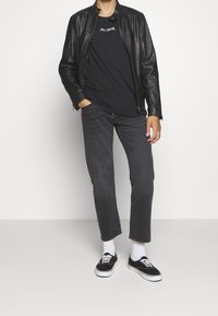 Schwarze Lederjacke, schwarzes T-Shirt mit Logo, graue Jeans und schwarze Sneakers mit weißen Schnürsenkeln. Socken sind über den Schuhen sichtbar.