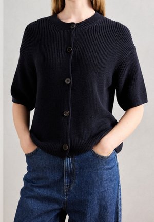 Vrouw draagt een marineblauw gebreid vest met korte mouwen en vijf knoppen, gecombineerd met blauwe spijkerbroeken, handen in de zakken.