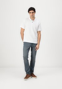 Hvid polo-shirt med krave og to sorte knapper, parret med lyseblå jeans og brune sneakers. Simpelt design og afslappet pasform.