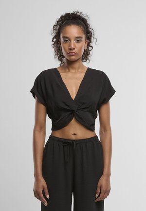 Vrouw met krullend haar in een hoge paardenstaart, draagt een zwart geknoopt cropped top en zwarte broek met trekkoord, staand tegen een effen achtergrond.