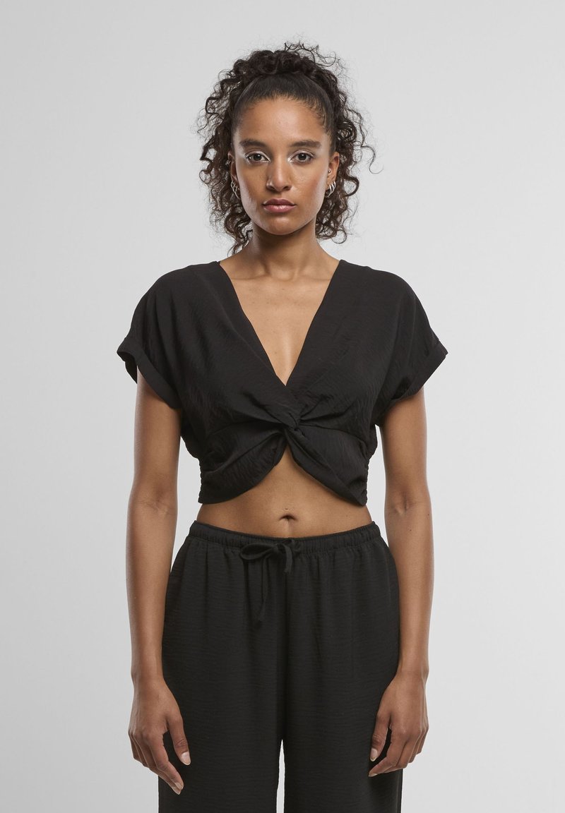 Femme aux cheveux bouclés en queue de cheval haute portant un crop top noir noué et un pantalon noir à cordon, debout devant un fond uni.