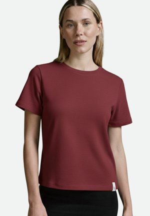 Bordeauxfarbenes Kurzarm-T-Shirt aus strukturiertem Stoff mit rundem Halsausschnitt. Verfügt über ein kleines Etikett am Saum und wird mit einem schwarzen strukturierten Rock kombiniert.