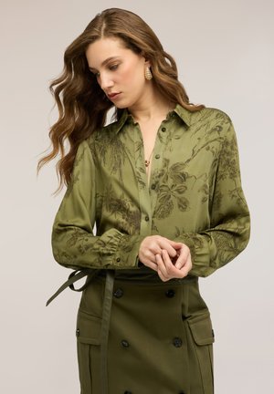 Kobieta o długich falujących włosach, ubrana w oliwkowo-zieloną bluzkę zapinaną na guziki z kwiatowym wzorem oraz dopasowaną spódnicę z czarnymi guzikami i kieszeniami.