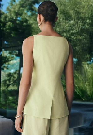 Naine kiiresti kokku pandud soenguga, kandes varrukateta helekollast topi ja sobivaid pükse, seisab siseruumides, kus on nähtav roheline välisvaade.