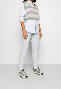 Gebreid vest in crème met lavendel- en groene hartpatronen over een lichtblauw button-up shirt, gecombineerd met licht denim spijkerbroeken en sneakers met een patroon.