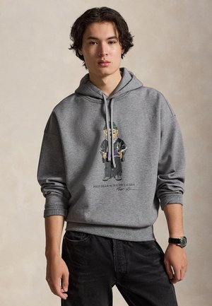 Un homme porte un sweat-shirt gris à capuche orné d’un motif Polo Bear et un jean noir, sur un fond beige uni.