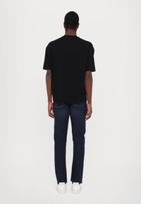 Černé tričko s krátkým rukávem a volným střihem, spárované s tmavě modrými džínami a bílými teniskami, z pohledu zezadu na jednobarevném pozadí.