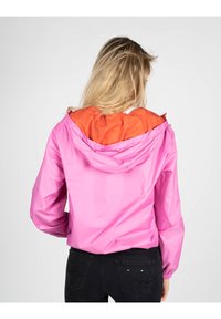 Giacca leggera rosa con cappuccio arancione, dotata di polsini elasticizzati e dettagli con coulisse. Indossata sopra jeans neri.