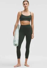 Sujetador deportivo de color verde oscuro y leggings de cintura alta, ambos hechos de tela elástica. La modelo sostiene una esterilla de yoga enrollada de color verde claro. Descalza.