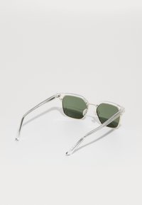 Ochelari de soare cu ramă transparentă, lentile verzi, formă geometrică și accente din metal pe balamale. Temele sunt transparente, cu detalii texturate.