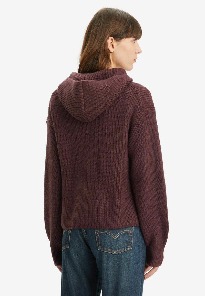 Bordeauxroter Strickpullover mit Kapuze, gerippter Struktur und langen Ärmeln. Kombiniert mit blauen Jeans, die einen entspannten Sitz und lässigen Stil zeigen.