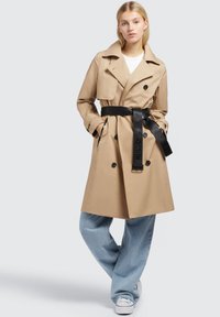 Trench coat bej cu un design cu dublu nasture, revere late, curea neagră și buzunare laterale, asociat cu blugi bleu deschis și adidași albi.