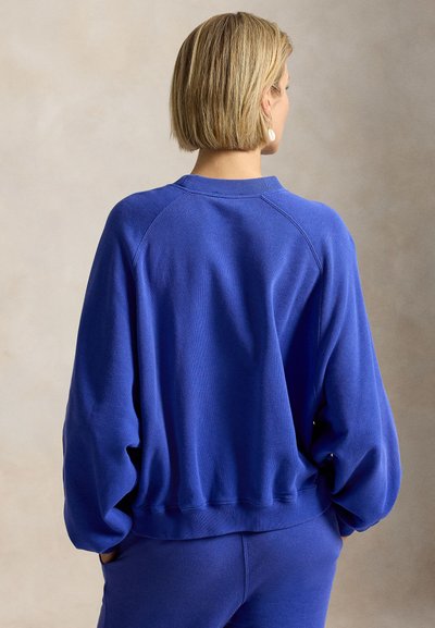 Γυναίκα με κοντά ξανθά μαλλιά που φοράει φαρδιά μπλε μπλούζα και παντελόνι σετ, όρθια με τα χέρια στις τσέπες, στραμμένη προς τα πίσω.