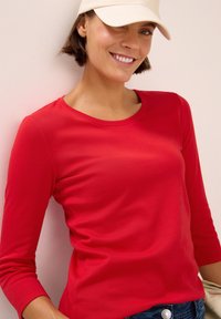 Rotes Langarmshirt mit rundem Halsausschnitt, aus weichem Stoff. Modell trägt eine beige Mütze, vor einem hellen Hintergrund.