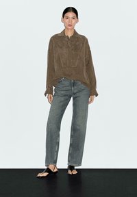 Camicia in suede marrone a maniche lunghe con colletto e patta anteriore, abbinata a jeans grigio chiaro a vita alta e sandali piatti neri.