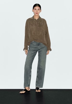 Camicia in suede marrone a maniche lunghe con colletto e patta anteriore, abbinata a jeans grigio chiaro a vita alta e sandali piatti neri.