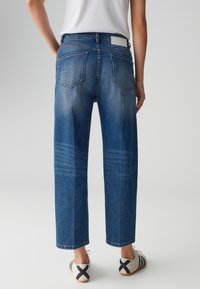 Jeans en denim bleu à coupe droite, avec poches arrière, légère décoloration et un patch de marque à la taille. Des chaussures décontractées complètent le look.