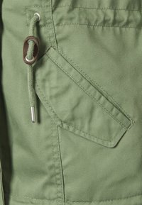 Grönt tyg med en slät textur, med en sydd ficka med en diagonal flik och en dragsko med metalldetaljer.