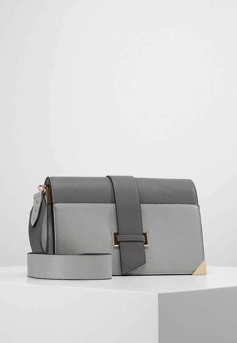 Szarawo-czarna prostokątna torba na ramię z szerokim paskiem, przednią klapką, złotym detalem na rogu i złotą klamrą, umieszczona na białej powierzchni.