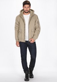 Beige pufferjakke med hætte, lynlås foran, to forlommer og ribbede manchetter. Bæret med en hvid T-shirt og mørke jeans, parret med sorte støvler.