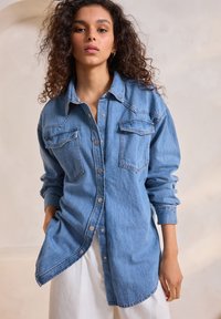 Camicia di jeans azzurro chiaro con maniche lunghe, due tasche frontali, orlo curvo e chiusura con bottoni a pressione. Tessuto testurizzato con dettagli cuciti.