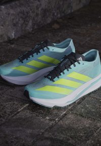 Jasnoniebieskie buty do biegania z czarnymi sznurówkami i trzema neonowymi żółtymi paskami. Gładka faktura z wyściełaną białą podeszwą i czarnymi akcentami.