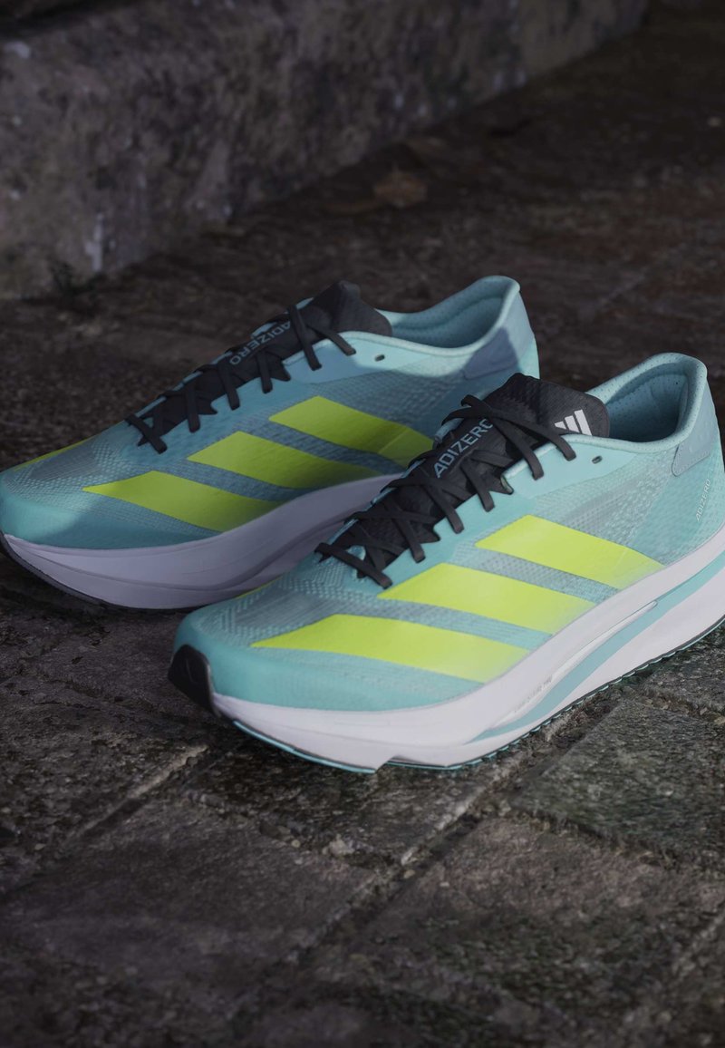 Zapatillas de running azul claro con cordones negros y tres rayas amarillas neón. Textura suave con una suela blanca acolchada y acentos negros.