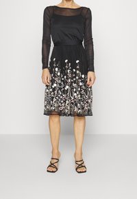 Frau in einem schwarzen, durchsichtigen langen Ärmeloberteil und einem schwarzen Rock mit mehrfarbiger Blumenstickerei, kombiniert mit schwarzen Riemchensandalen.