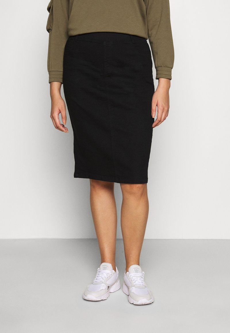 Femme portant une jupe crayon noire longueur genou, un haut à manches longues vert olive, et des baskets blanches, debout devant un fond uni.