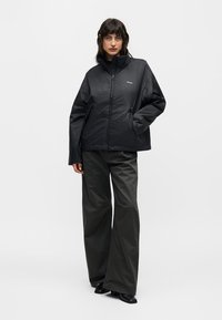 Γυναίκα όρθια, φοράει μαύρο μπουφάν με φερμουάρ και ψηλό γιακά και φαρδιά σκούρα παντελόνια, με φόντο απλό ανοιχτό χρώμα.