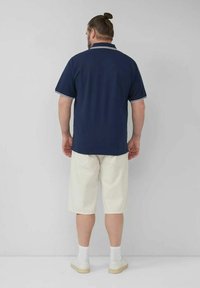 Navyblaues Poloshirt mit weißem Saum, kombiniert mit hellbeigen Shorts. Glattes Material, normale Passform, Rückansicht, präsentiert mit weißen Socken und Schuhen.