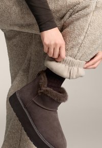 Rjavi semiš čevlji do gležnja z obrobo iz krzna, kombinirani z bež flis hlačami in temnimi rebrastimi nogavicami, ki poudarjajo teksturirane materiale in sproščen kroj.