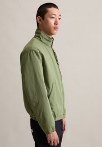 Jonge man in een groene blouson met hoge kraag staat in profiel tegen een effen beige achtergrond.