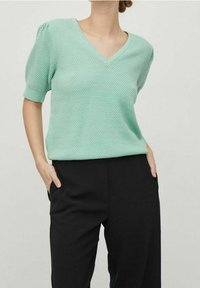 Top lavorato a maglia verde menta con scollo a V e maniche corte a sbuffo, abbinato a pantaloni neri, che presenta un design testurizzato e una vestibilità rilassata.