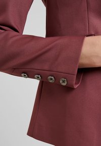Rukáv burgundského blejzru s hladkou texturou, zakončený pěti stříbrnými knoflíky s kruhovými vzory a upraveným střihem.