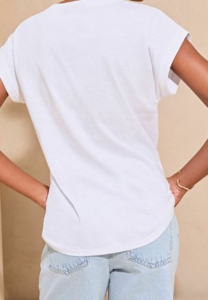 Persona con una camiseta blanca de manga corta lisa y jeans de mezclilla azul claros, de pie con las manos en las caderas contra un fondo beige.