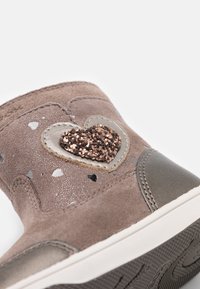 Hnědá semišová dětská bota se třpytivou srdíčkovou nášivkou a bílými a šedými puntíky, detailní záběr ukazuje texturovanou podrážku a prošívání.