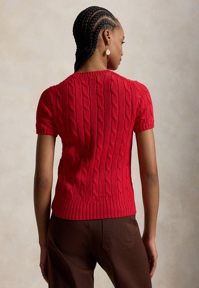 Kobieta z warkoczami, ubrana w czerwony sweter warkoczowy z krótkim rękawem i brązowe spodnie, widziana od tyłu na neutralnym tle.