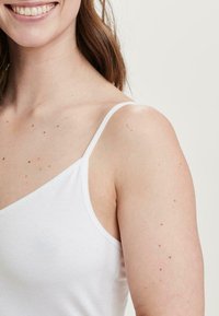 Hvid bomulds tanktop med tynde stropper, der har en V-udskæring og en tætsiddende design. Glat tekstur, der fremhæver hudfarve og lette fregner.