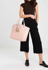 Femme portant un pantalon noir raccourci et un haut marron à manches courtes, tenant un grand sac fourre-tout rose clair avec des détails à pompons.
