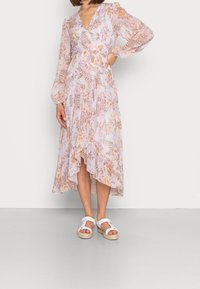 Lång blommig klänning i ljuslila med persiko- och gröna nyanser, med en omslagsdesign, flödande nederdel och genomskinliga, texturerade ärmar. Bärs med vita sandaler.