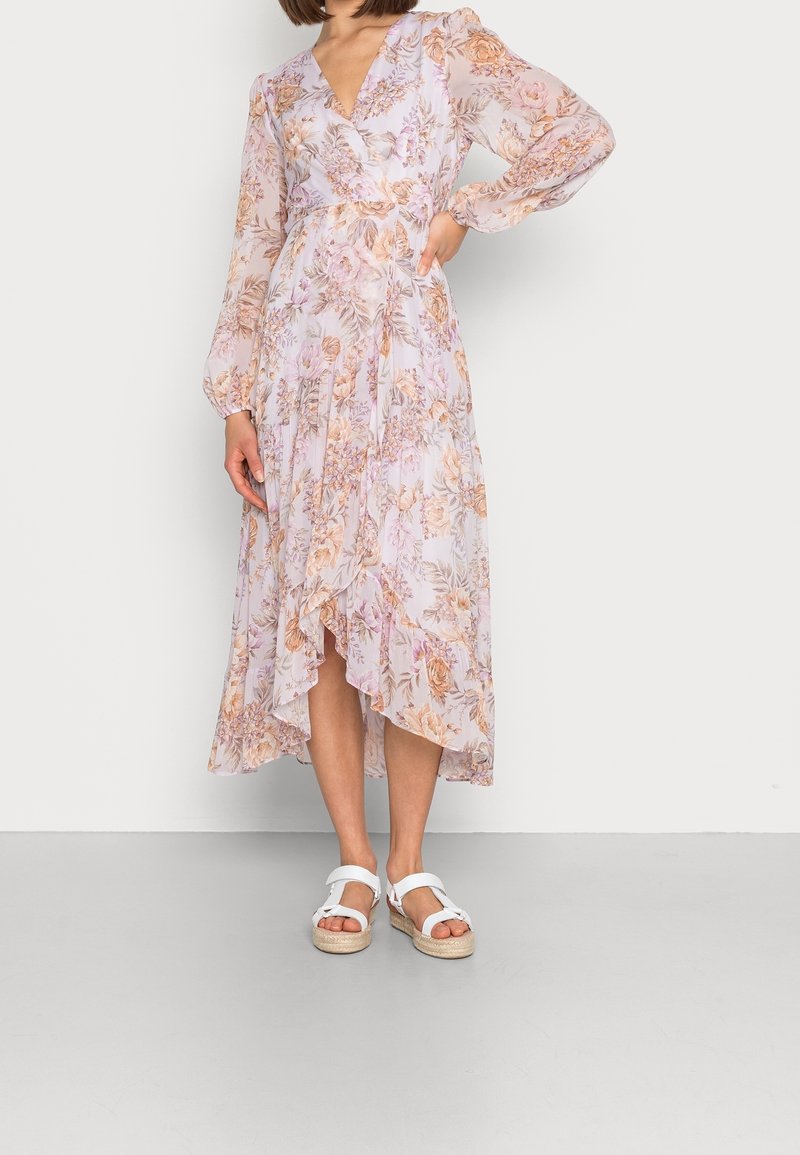 Lång blommig klänning i ljuslila med persiko- och gröna nyanser, med en omslagsdesign, flödande nederdel och genomskinliga, texturerade ärmar. Bärs med vita sandaler.