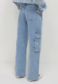 Persona che indossa pantaloni cargo in denim azzurro chiaro e una giacca in denim coordinata, in piedi con le mani nelle tasche davanti a uno sfondo neutro.