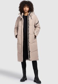Beige pufferjacka med huva, med dragkedja fram, tryckknappar och sidofickor. Svarta detaljer och logotyp finns.