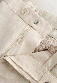 Pantalon beige sur mesure avec fermeture par bouton, crochet en métal et fermeture éclair, présentant des coutures détaillées et une doublure intérieure à motif.