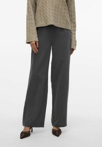 Spodnie o szerokich nogawkach w ciemnoszarym materiale, charakteryzujące się gładką fakturą i dopasowanym krojem. Połączone z jasnobeżowym swetrem w splot warkoczowy i ciemnymi szpilkami.