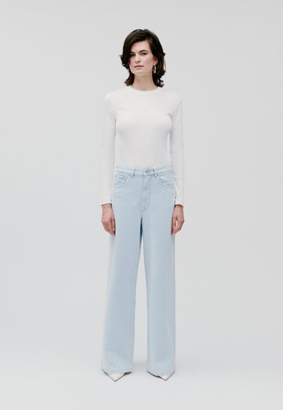 Jeans a gamba larga blu chiaro con vita alta, abbinati a un top fitted bianco a maniche lunghe. Tessuto liscio con un design minimale e pulito.