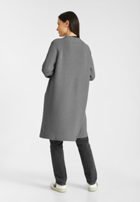 Grijze geribde cardigan met een lange, losse pasvorm, voorzien van een sjawlboord en zijsplitten. Gepaarde met donkere jeans en witte sneakers.